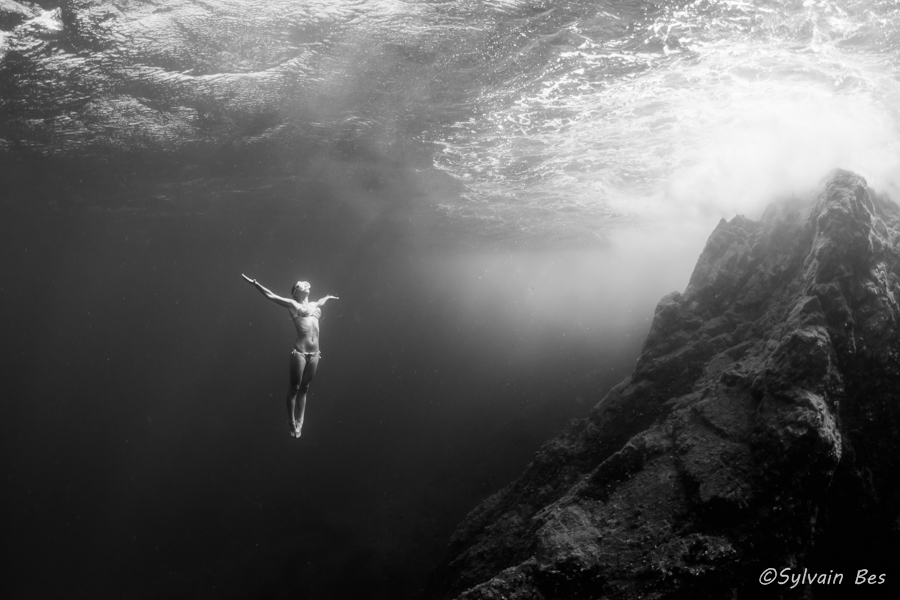 Une femme en bikini et en apnée regagne la surface les bras en croix. La photographie est en noir et blanc. A gauche la femme et à droite on aperçoit des rochers sous-marins